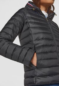 Veste matelassée noire avec fermeture éclair devant, poches latérales zippées et capuche bordée de rayures rouges, blanches et bleues.