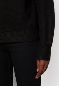 Pull en maille noire avec des poignets côtelés, assorti à un pantalon noir. Présente un petit logo rouge, blanc et bleu sur la manche gauche.