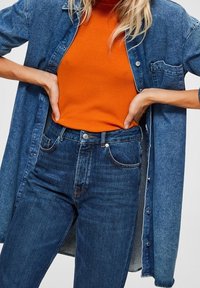 Denimskjorta med knappar, bärs öppen över en ribbad orange topp. Höga mörkblå jeans med framfickor och lätt slitning.