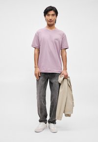T-shirt lavanda a maniche corte con taschino frontale, abbinato a jeans grigi strappati. Il modello tiene in mano una giacca beige e indossa sneakers bianche.