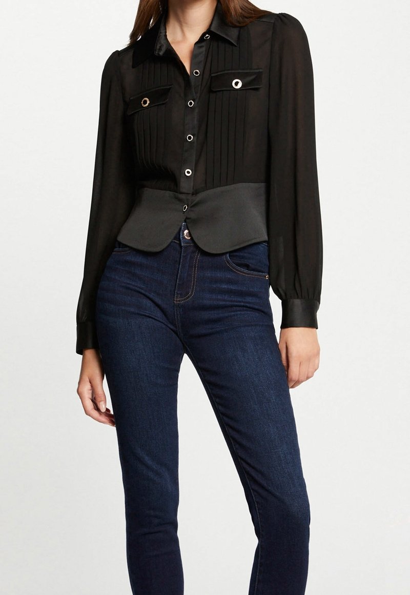Femme portant un chemisier noir transparent boutonné avec des plis et deux poches frontales, associé à un jean bleu foncé ajusté.