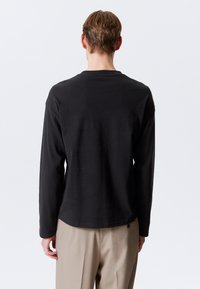 Tricou negru cu mânecă lungă, având un guler rotund și o croială lejeră. Materialul pare neted, cu un design simplu, purtat peste pantaloni bej.