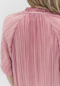 Pleated, vaaleanpunainen paita, jossa on pehmeä rakenne ja pyöreä pääntie. Takana on hopeinen lenkkikoriste ja lyhyet, löysät hihat.