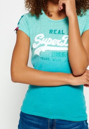 Personne portant un t-shirt SuperDry bleu-vert, bras croisés et une main reposant sur le menton, visible du cou aux hanches.