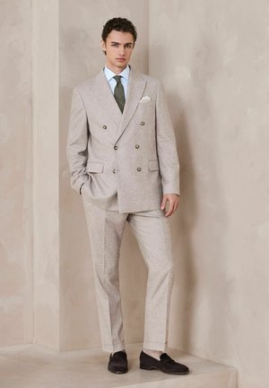 Junger Mann in einem beigen, doppelt geknöpften Anzug, grüner Krawatte, hellblauem Hemd und schwarzen Loafers vor einer strukturierten beigen Wand.