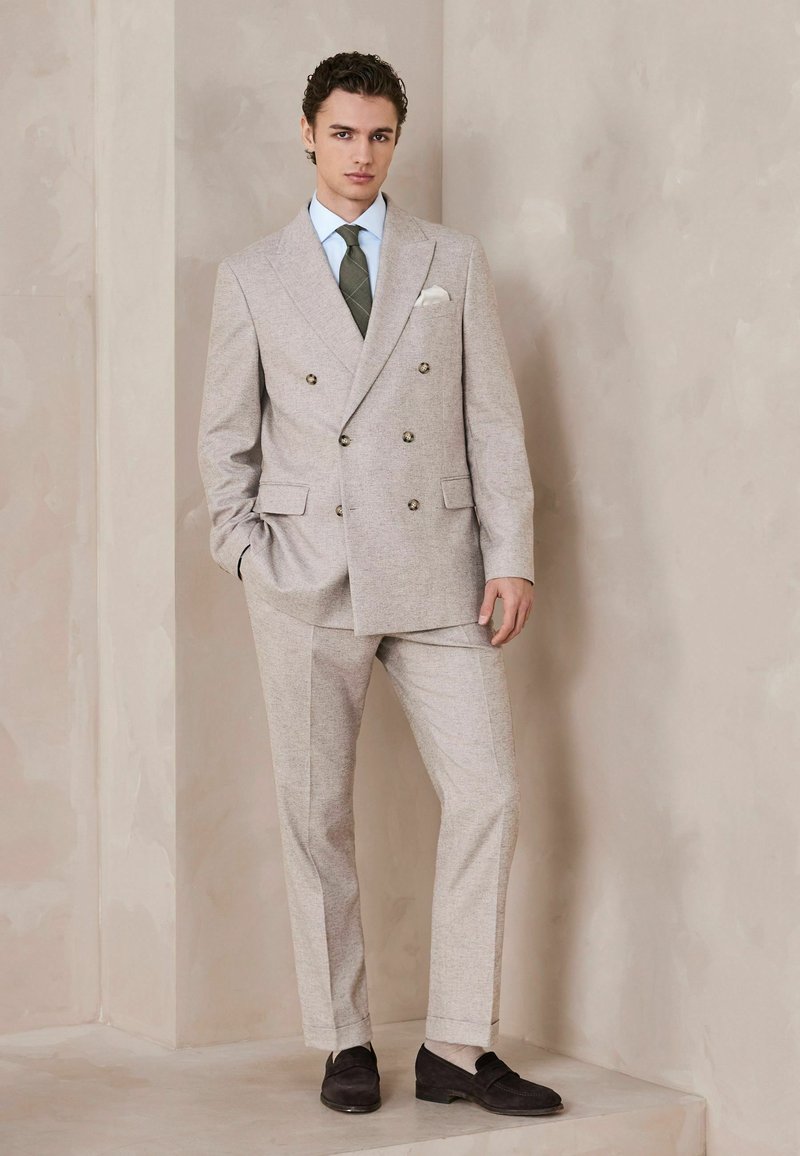 Mladý muž stojaci v béžovom dvojradovom obleku, so zelenou kravatou, svetlomodrou košeľou a čiernymi mokasínami pred textúrovanou béžovou stenou.