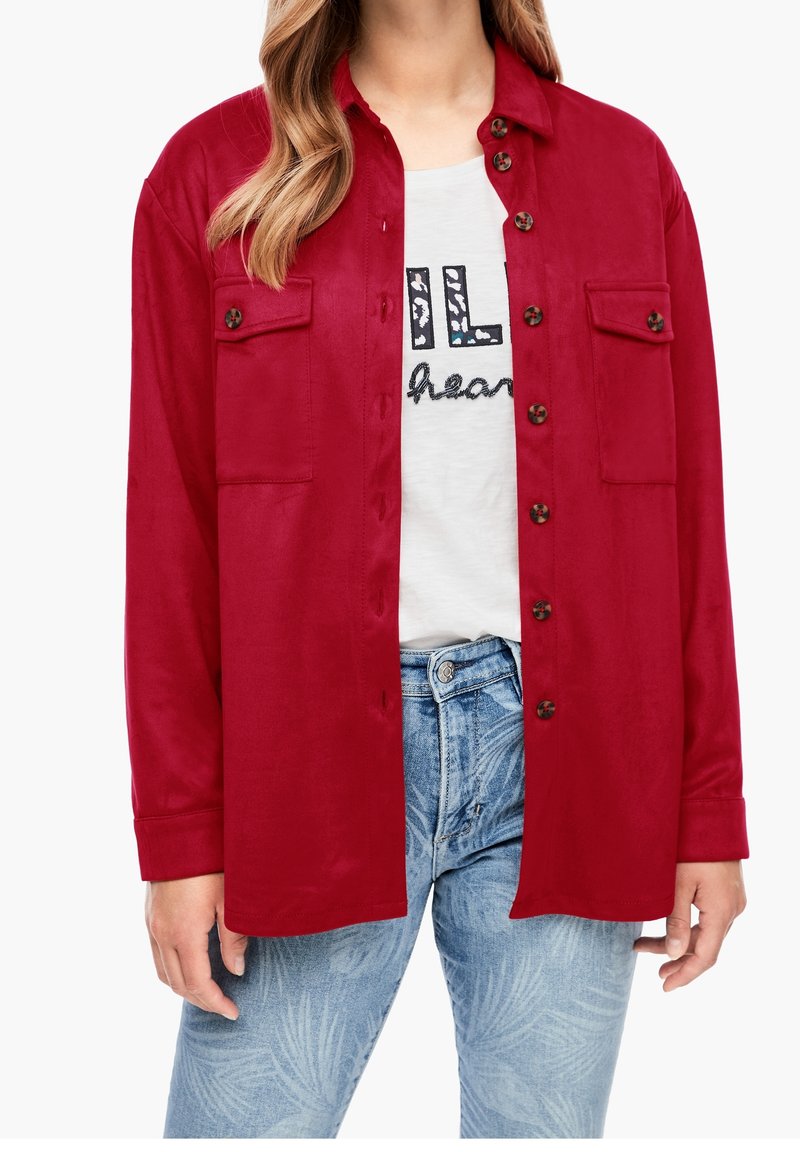 Kvinde iført en rød knappet skjorte over en hvid t-shirt med grafik og lyseblå jeans med bladmønster.