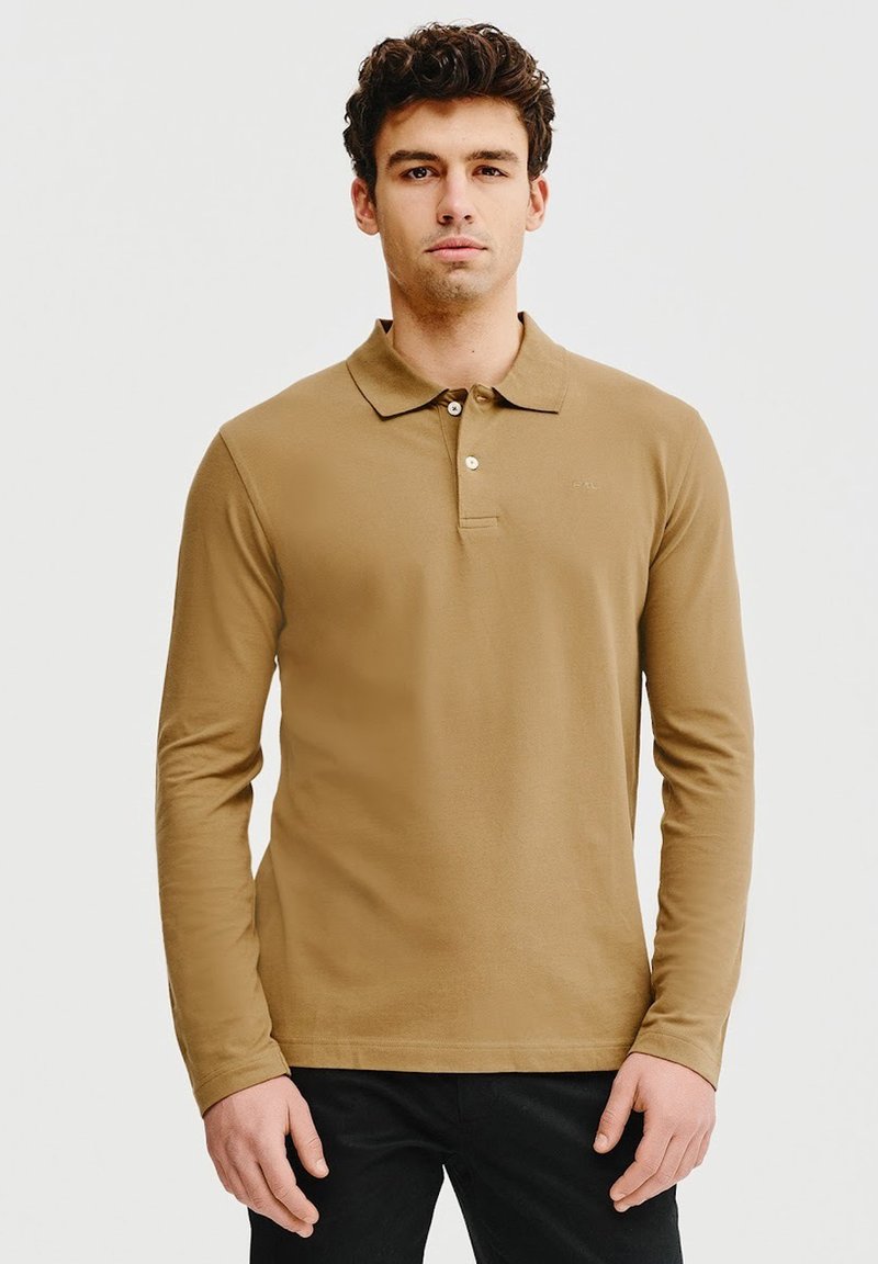 Beżowa polo z długim rękawem i kołnierzykiem, z trzema guzikami i miękkim materiałem. Model zestawiony z czarnymi spodniami.