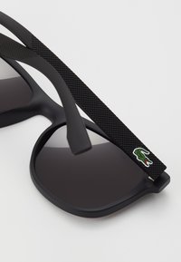 Ochelari de soare negri cu lentile întunecate. Brațe texturate negre cu un mic logo verde de crocodil. Design cu ramă rotundă.