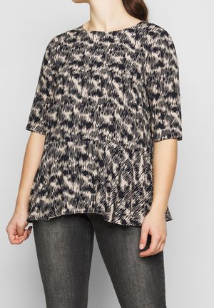 Femme portant un haut péplum à manches courtes avec un motif abstrait noir et beige, associé à un jean gris foncé taille slim.