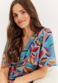 Květinové šaty z modré látky s barevnými vzory listů. Krátké rukávy, výstřih do V a nařasený detail na poprsí. Měkká textura.