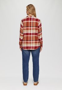 Rood en beige geruite overhemd met lange mouwen en knoopmanchetten, met een licht gebogen zoom, gedragen met blauwe jeans en lichtgekleurde schoenen.