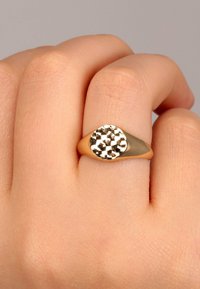 Guldtonad ring med en rund, fasetterad metalltopp. Slät ring med en polerad finish, bärs på en hand med ljus hudton.