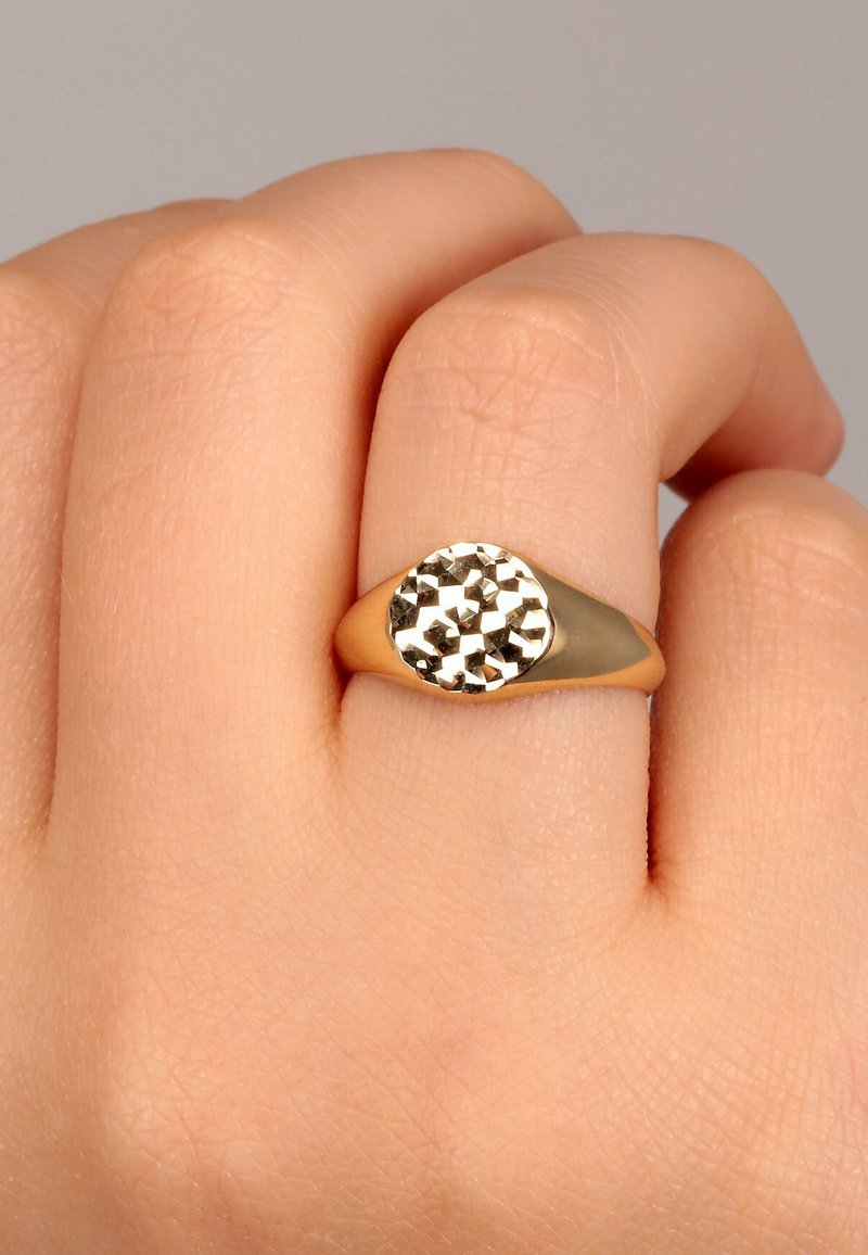 Guldtonad ring med en rund, fasetterad metalltopp. Slät ring med en polerad finish, bärs på en hand med ljus hudton.