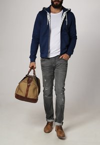 Marinblå zip-up hoodie med vita snören, vit t-shirt, slitna grå jeans, bruna snörskor och en beige duffelväska med bruna detaljer.