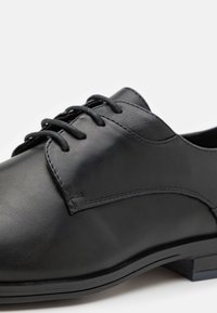 Chaussure habillée noire en cuir à lacets avec des panneaux lisses et texturés et un talon bas sur fond blanc.