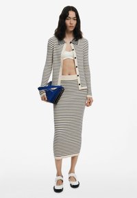 Set di cardigan e gonna a maglia a righe in crema e blu navy, con bottoni, texture a coste e silhouette aderente. Il modello tiene in mano una borsa blu.