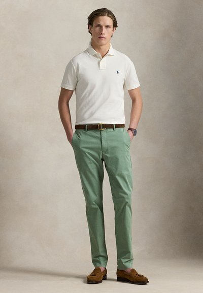 Ung mann står med hendene i lomma, iført hvit poloskjorte, grønne bukser, brun belte, klokke og brune loafers foran en ensfarget bakgrunn.