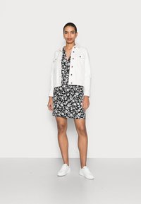 Jaqueta de ganga branca sobre um vestido floral preto, com decote em V e mangas curtas. Combinada com ténis brancos, em pé sobre um chão cinza.