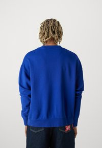 Felpa blu con collo a giro, vestibilità rilassata, polsini e orlo a coste. Tessuto morbido; la vista da dietro mostra una trama liscia, senza decorazioni.