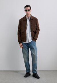 Uomo con occhiali da sole neri, giacca in suede marrone, camicia bianca con bottoni, jeans blu e scarpe nere, in piedi contro un muro semplice.