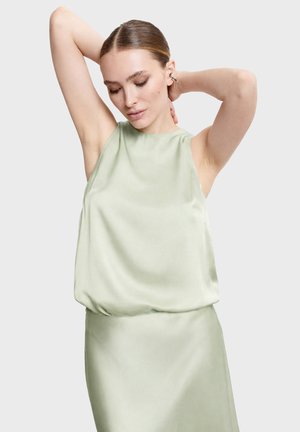 Žena so vzadu uhladenými vlasmi, oblečená v svetlozelenom saténovom topánku bez rukávov a sukni, pózujúca s rukami zdvihnutými za hlavou.