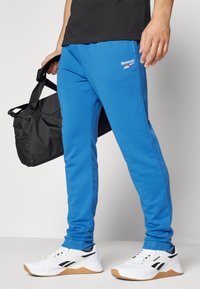 Zili sporta bikses ar ribotām malām, ar Reebok logo. Melna sporta soma turēta rokās; valkātas baltas kurpes ar melniem akcentiem.