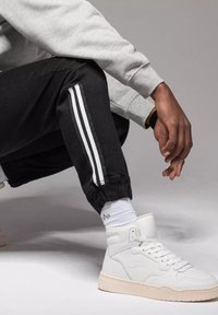 Czarne dresowe spodnie z białymi paskami po bokach, elastyczne mankiety, w połączeniu z białymi sneakersami za kostkę i szarym swetrem. Tło o podłożu ziemnym.