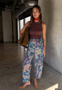 Γυναίκα που στέκεται μέσα σε κτίριο, φορώντας παντελόνι με μοτίβο παϊέντζα, αμάνικο σκούρο τοπ, κόκκινο μαντήλι στο λαιμό, κρατώντας μεγάλη καφέ τσάντα σουέντ και καφέ μυτερά παπούτσια.