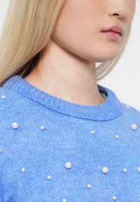Młoda osoba z długimi blond włosami, ubrana w niebieski sweter dzianinowy ozdobiony małymi białymi perłami na białym tle.