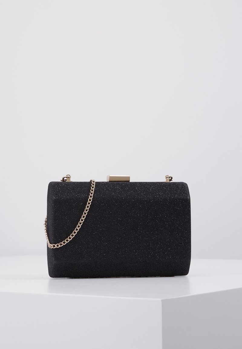 Pochette rectangulaire noire avec texture fine pailletée et bandoulière chaîne dorée, exposée sur une surface blanche devant un fond gris.