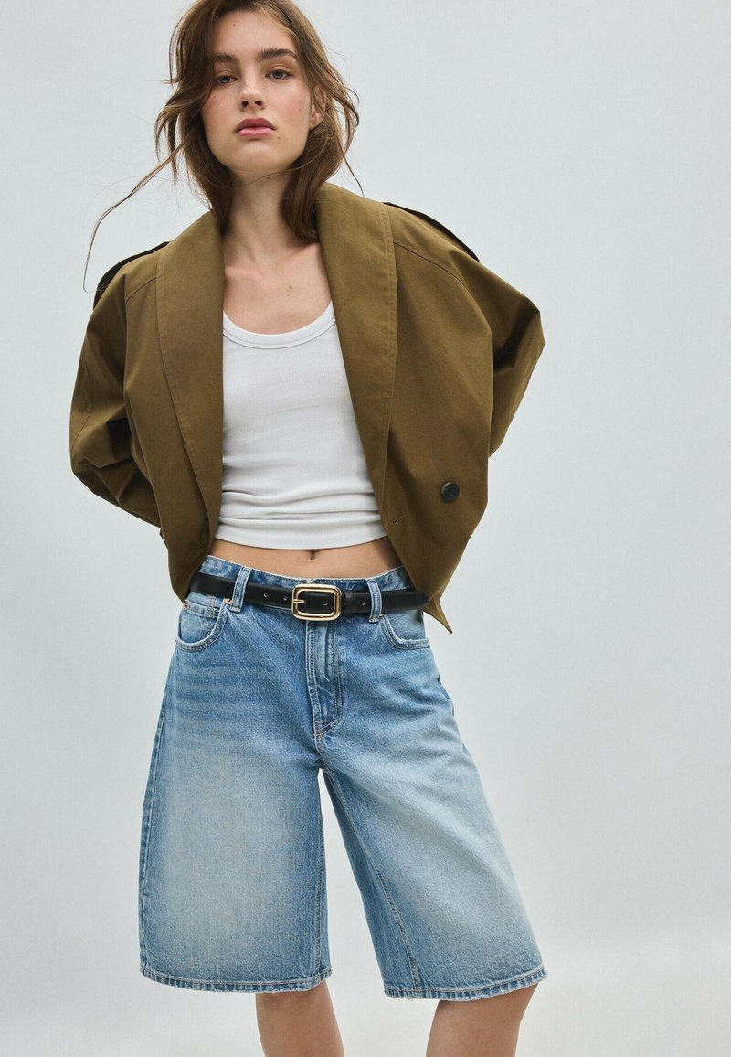 Jeune femme portant une veste courte olive, un débardeur blanc et un short bermuda en denim bleu clair avec une ceinture noire, se tenant devant un fond uni.