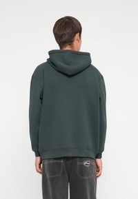 Person wearing dark green hooded sweatshirt and black jeans, facing away against a plain white background.