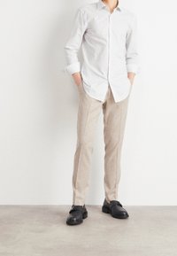 Persoon in beige broek, een wit gestreept overhemd met opgerolde mouwen, zwarte instappers, staande tegen een eenvoudige witte muur.