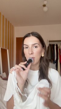 Modelis laiko juodą makiažo šepetėlį arti burnos, laikydamas baltą drabužį su nėriniuota apdaila. Kambaryje minimalus interjeras ir dryžuotos sienos.