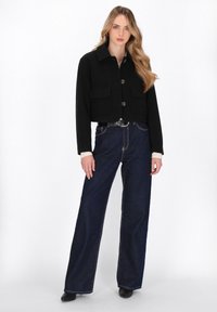 Kvinde med langt blondt hår iført en sort cropped jakke, brede mørkeblå jeans, sort bælte og sorte spidse sko, stående foran en hvid baggrund.