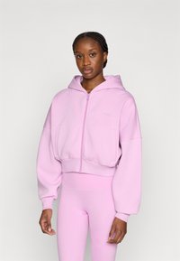 Ροζ κοντό φούτερ από μαλακό ύφασμα, με φερμουάρ μπροστά, μεγάλο κουκούλι και φαρδιά μανίκια, σε συνδυασμό με ροζ κολάν που ταιριάζουν.