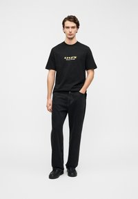 Μαύρο βαμβακερό t-shirt με κίτρινο λογότυπο, σε συνδυασμό με φαρδιά μαύρα τζιν και μαύρα παπούτσια, που στέκεται μπροστά σε απλό φόντο.