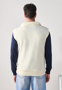 Fleece vest i creme, med en bred krave og ærmeløs design. Båret over en marineblå langærmet skjorte, parret med lyseblå jeans.