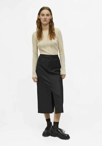 Mlada ženska z dolgimi lasmi nosi kremast pleten top, črno midi krilo s sprednjim razporkom, črne nogavice in deležne črne vezalne čevlje na debelo, v obrazcu pred belo ozadje.