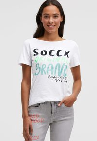 Weißes T-Shirt mit kurzen Ärmeln und mehrfarbigem Text: "SOCCX ORIGINAL BRAND" und "Kap Verde." Kombiniert mit hellgrauen Jeans mit Print-Details.
