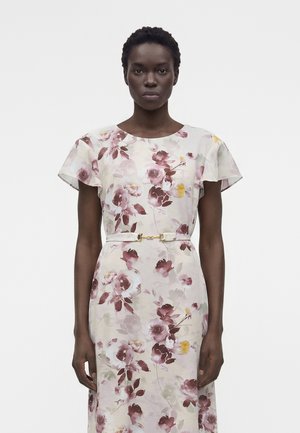 Femme aux cheveux courts et bouclés, portant une robe claire avec des motifs floraux roses et violets et une fine ceinture blanche avec une boucle dorée.