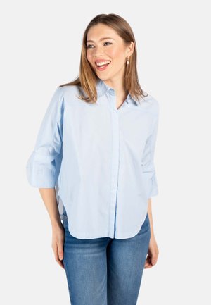 Ung kvinne smilende, iført lyseblå skjorte med knapper og trekvartlange ermer og blå jeans, stående foran hvit bakgrunn.