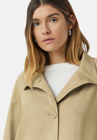 Beżowa kurtka bez kołnierza z zapięciem na guziki, charakteryzująca się gładkim materiałem i minimalistycznym designem. Dyskretne detale przeszycia w okolicy kołnierza.