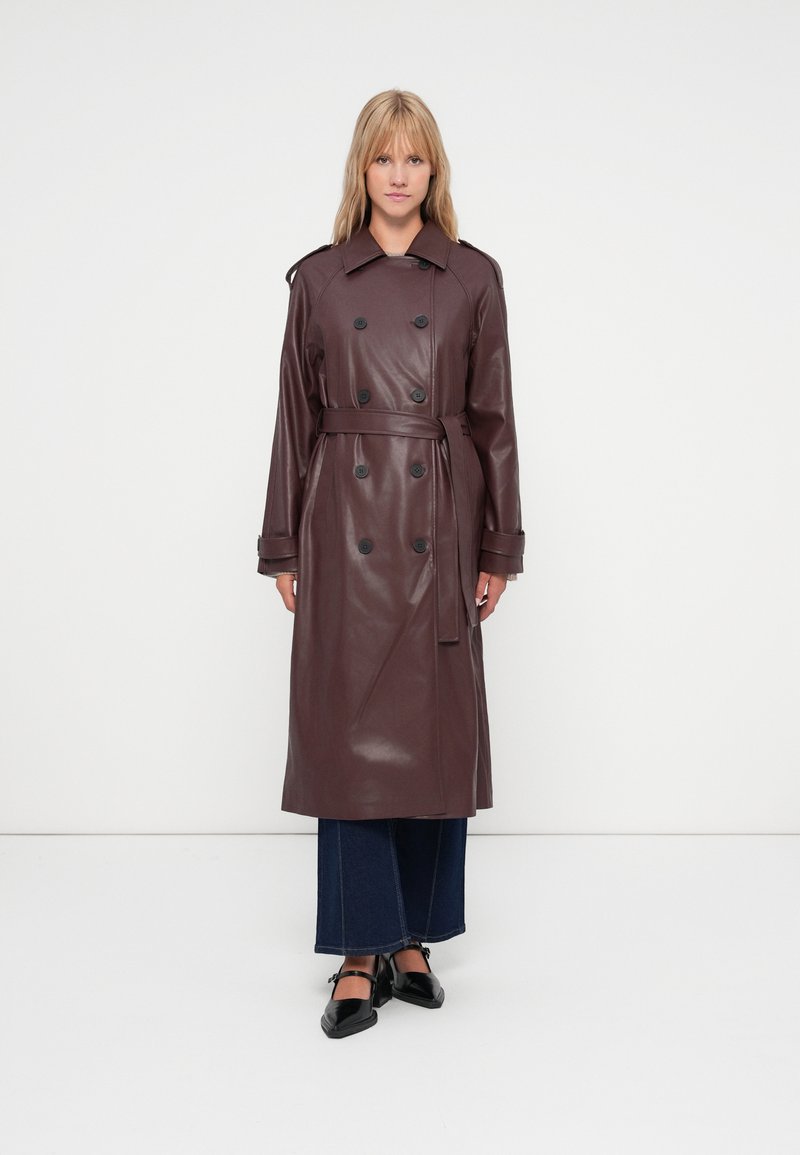 Burgundy kein nahkaa oleva trench-coat, jossa on kaksirivinen muotoilu, mustat napit, leveä kaulus ja vyö vyötäröllä. Käytetty leveälahkeisten farkkujen päällä.