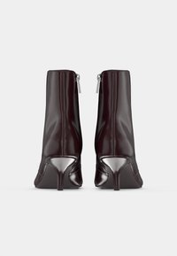 Pár lesklých tmavohnedých členkových čižiem s úzkymi vysokými podpätkami a viditeľnými zadnými zipsami, zobrazených zozadu na čistej bielej pozadí.