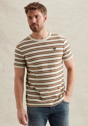 Mężczyzna ubrany w beżowo-brązowo-białą koszulkę w paski z krótkim rękawem i małym czarnym emblem na lewej piersi, w połączeniu z niebieskimi dżinsami.