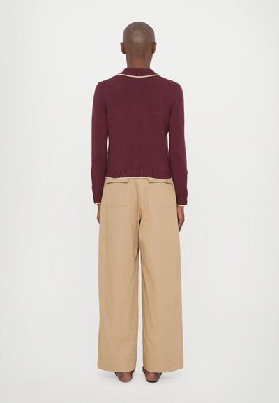 Pull en maille bordeaux avec un ourlet et des poignets côtelés, présentant un subtil liseré jaune. Associé à un pantalon beige large.
