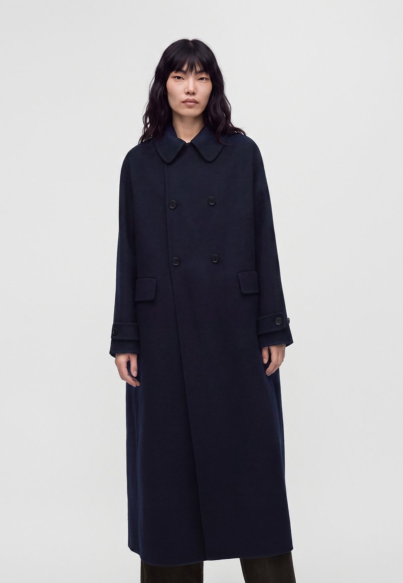 Femme aux longs cheveux noirs portant un long manteau sombre à double boutonnage avec un large col, debout devant un fond clair uni.
