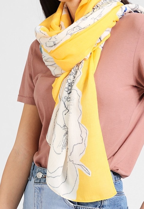 Femme portant une écharpe fleurie jaune et blanche sur un haut à manches courtes rose poudré et un jean bleu clair, nouée de façon lâche.